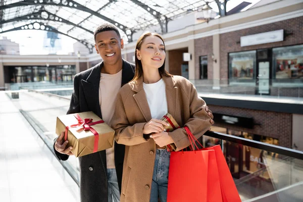 Feliz e elegante casal interracial com caixas de presente e sacos de compras perto do shopping borrado — Fotografia de Stock