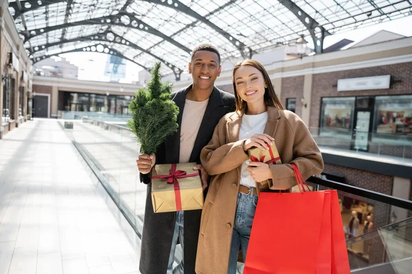 Elegante pareja multiétnica sosteniendo bolsas de compras y pequeño árbol de Navidad con cajas de regalo cerca del centro comercial - foto de stock