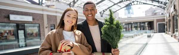 Pareja interracial de moda con cajas de regalo y pequeño árbol de Navidad sonriendo cerca del centro comercial borroso, pancarta - foto de stock