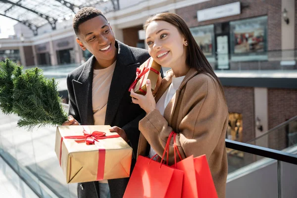 Curiosa mujer sacudiendo caja de regalo cerca de hombre afroamericano con regalos y pequeño árbol de Navidad - foto de stock