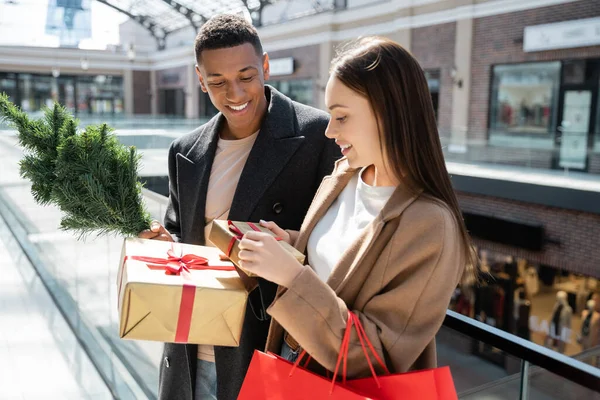 Femme excitée avec des sacs à provisions tenant boîte cadeau près souriant homme afro-américain avec petit arbre de Noël — Photo de stock