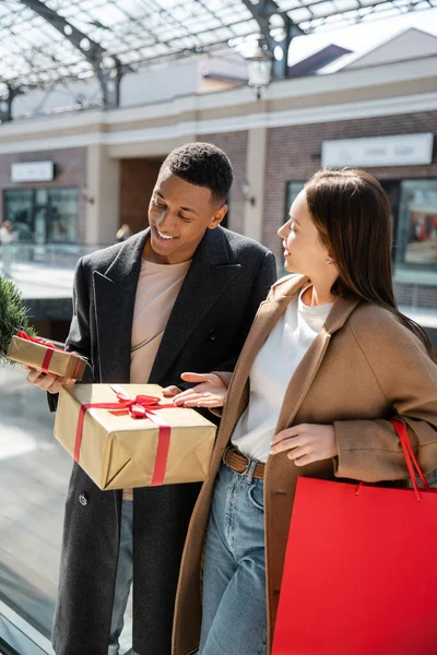 Homem americano africano alegre segurando caixas de presente perto de jovem namorada com sacos de compras — Fotografia de Stock
