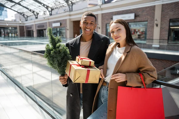 Pareja multiétnica de moda con regalos de año nuevo y pequeño árbol de Navidad sonriendo a la cámara cerca del centro comercial borroso - foto de stock