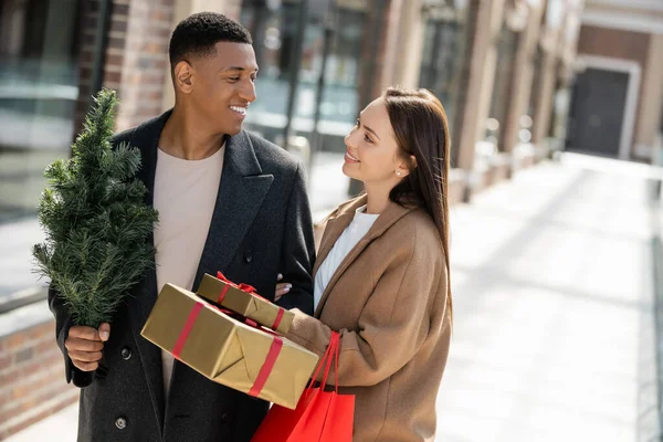 Счастливая женщина с пакетами для покупок улыбается африканскому американцу с подарочными коробками и елкой — стоковое фото