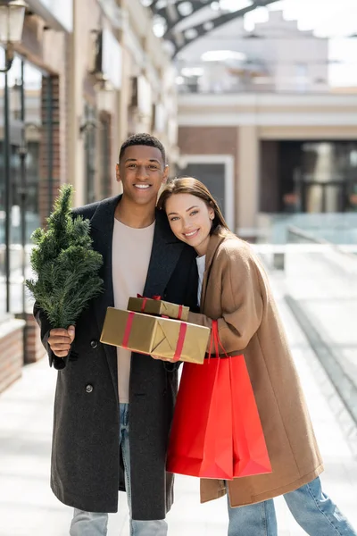 Pareja interracial de moda con bolsas de compras y cajas de regalo con pequeño árbol de Navidad sonriendo a la cámara en la calle - foto de stock