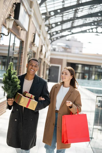 Happy and trendy interracial couple with small christmas tree and presents on blurred urban street — Stock Photo