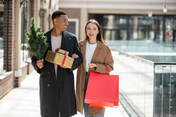 Hombre afroamericano con pequeño árbol de navidad y cajas de regalo mirando a la novia feliz con bolsas de compras - foto de stock