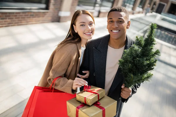 Feliz y de moda pareja multiétnica con bolsas de compras y regalos de año nuevo con pequeño árbol de Navidad en la calle de la ciudad - foto de stock