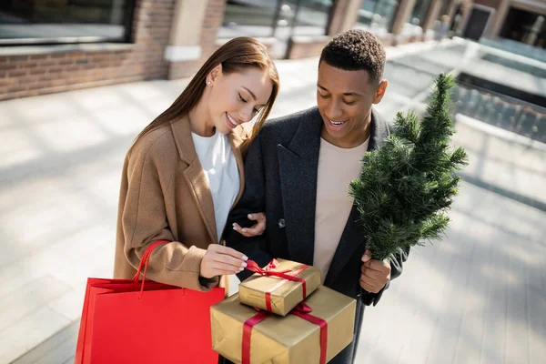 Mujer feliz con bolsas de compras mirando cajas de regalo cerca de hombre afroamericano con pequeño árbol de Navidad - foto de stock