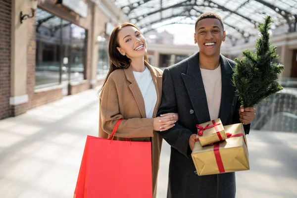 Fröhliches gemischtrassiges Paar mit Einkaufstaschen und Geschenkschachteln mit kleinem Weihnachtsbaum, der in die Kamera in der Nähe des Einkaufszentrums blickt — Stockfoto