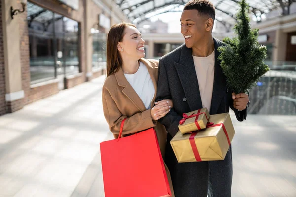 Élégant couple interracial en manteaux tenant de nouveaux cadeaux de l'année et petit arbre de Noël tout en se souriant — Photo de stock