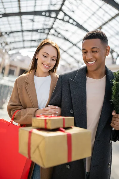 Alegre pareja multiétnica en abrigos de moda mirando borrosas cajas de regalo - foto de stock