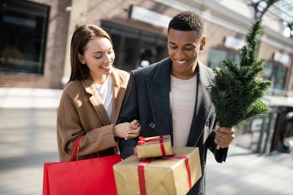 Mujer alegre con bolsas de compras mirando cajas de regalo cerca de hombre afroamericano con pequeño árbol de Navidad - foto de stock