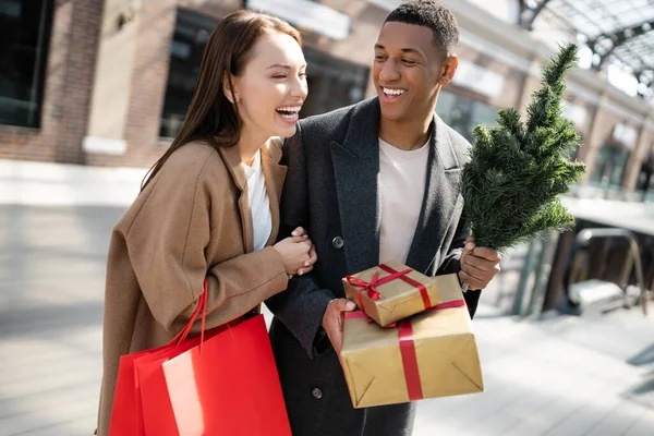 Mujer emocionada con bolsas de compras riendo cerca de hombre afroamericano elegante celebración de regalos y pequeño árbol de Navidad - foto de stock