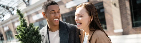 Young excited woman laughing near smiling african american man with small christmas tree, banner — Stock Photo