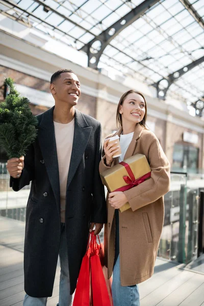 Elegante casal interracial com novos presentes de ano e pequena árvore de Natal sorrindo perto de shopping borrado — Fotografia de Stock