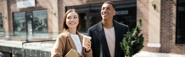 Mujer sonriente con café para llevar y caja de regalo mirando hacia otro lado cerca de hombre afroamericano con pequeño árbol de Navidad, pancarta - foto de stock