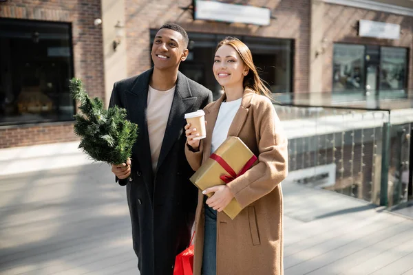 Giovane donna con caffè per andare e scatola regalo sorridente vicino alla moda afro-americano uomo con piccolo albero di Natale — Foto stock