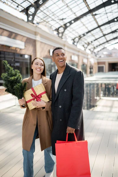 Счастливая и модная межрасовая пара держит сумку и подарочную коробку с рождественской елкой возле размытого торгового центра — стоковое фото