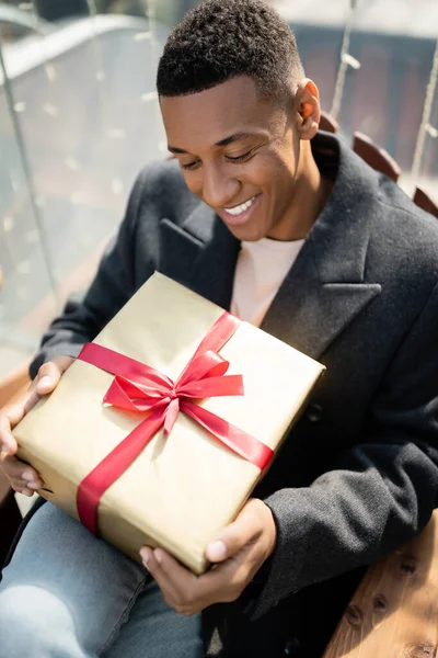 Счастливый африканский американец в пальто сидит с подарочной коробкой на открытом воздухе — стоковое фото