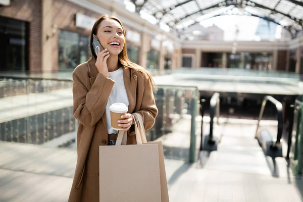 Excited woman talking on mobile phone while standing with shopping bags and coffee to go near blurred mall — Stock Photo