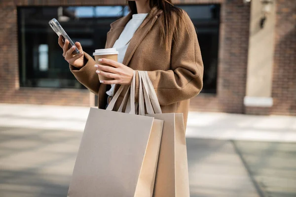 Ausgeschnittene Ansicht einer Frau, die ihr Smartphone benutzt und Einkaufstüten mit Pappbecher auf der Straße hält — Stockfoto