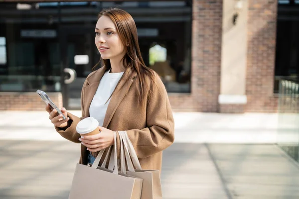 Donna sorridente in beige appendiabiti smartphone mentre in piedi con borse della spesa e tazza di carta su strada sfocata — Foto stock
