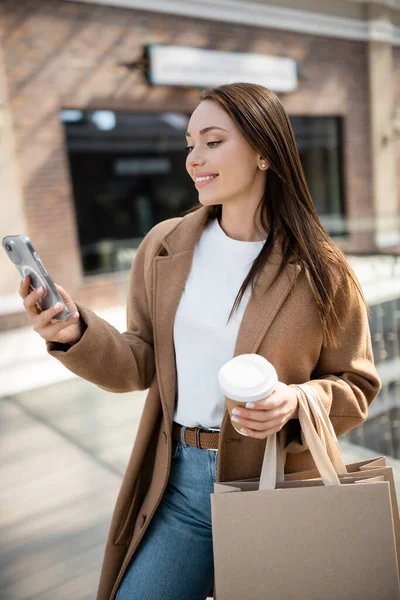 Glückliche brünette Frau im Mantel, die auf das Smartphone schaut und Einkaufstüten mit Pappbecher auf der Straße hält — Stockfoto