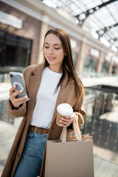 Trendy brunette woman holding paper cup and shopping bags while looking at smartphone — Stock Photo