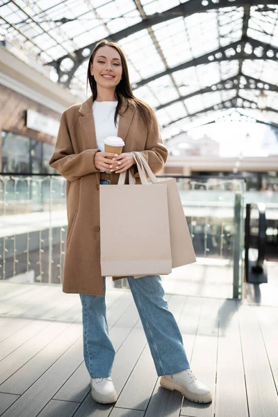 Pleine longueur de femme souriante en manteau et jeans debout avec des sacs à provisions et du café pour aller près du centre commercial flou — Photo de stock