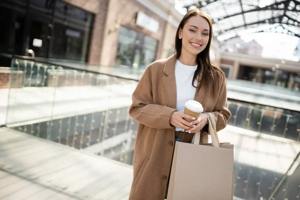 Mujer feliz y elegante con bebida para llevar y bolsas de compras mirando a la cámara cerca del centro comercial borroso - foto de stock