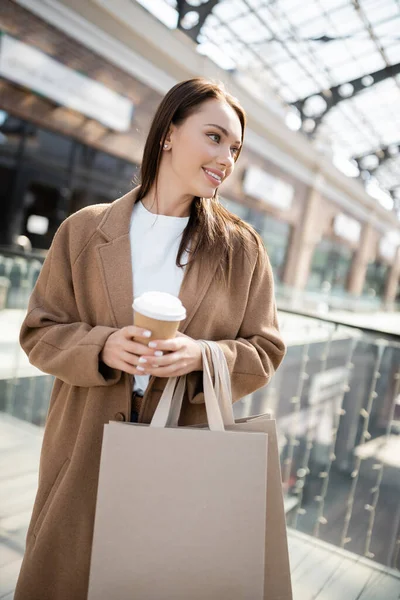 Femme souriante en manteau beige tenant sacs à provisions et tasse en papier tout en regardant loin près du centre commercial flou — Photo de stock