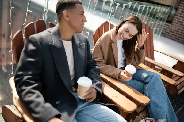 Cheerful interracial couple in trendy coats sitting with coffee to go on wooden seats on city street — Stock Photo