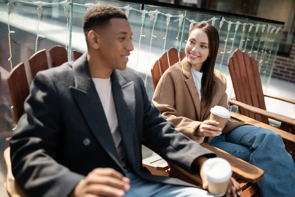 Sourire couple interracial en manteaux assis avec des tasses en papier sur des sièges en bois à l'extérieur — Photo de stock