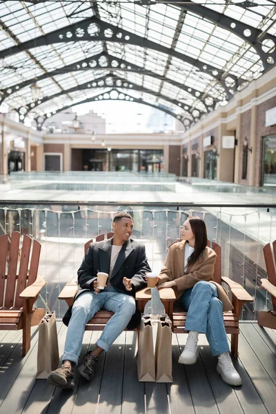 Homem americano africano na moda falando com a namorada enquanto sentado perto de sacos de compras e shopping borrado — Fotografia de Stock