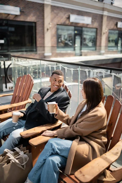 Alegre pareja multiétnica en abrigos sentados con café para ir cerca de bolsas de compras y centro comercial borrosa - foto de stock