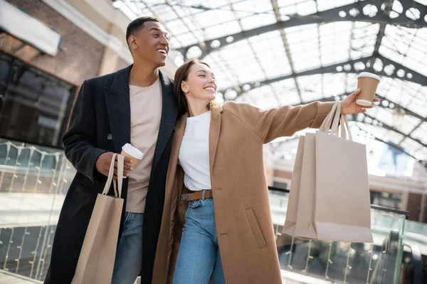Mulher na moda com copo de papel e sacos de compras apontando com a mão perto do namorado americano africano animado — Fotografia de Stock