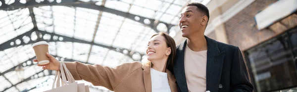 Lächelnde Frau mit Einkaufstaschen und Imbissgetränk, die mit der Hand in die Nähe eines erstaunten afrikanisch-amerikanischen Mannes zeigt, Banner — Stockfoto