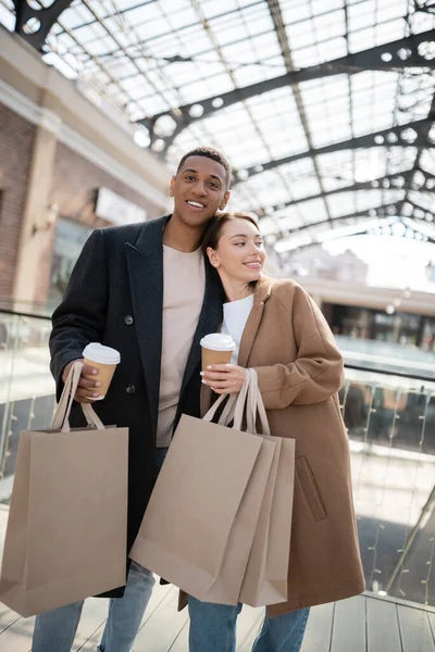 Felice ed elegante coppia multietnica con bicchieri di carta e borse della spesa sorridente vicino centro commerciale offuscata — Foto stock