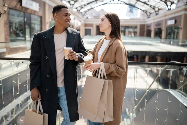 Stylish interracial couple with shopping bags and paper cups smiling at each other near blurred mall — Stock Photo