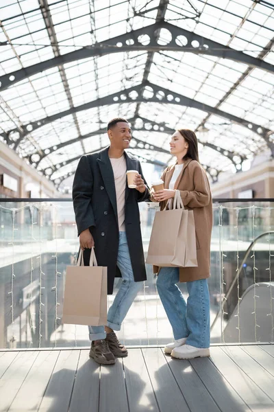 Full length of interracial couple in trendy coats standing with paper cups and purchases near mall — Stock Photo