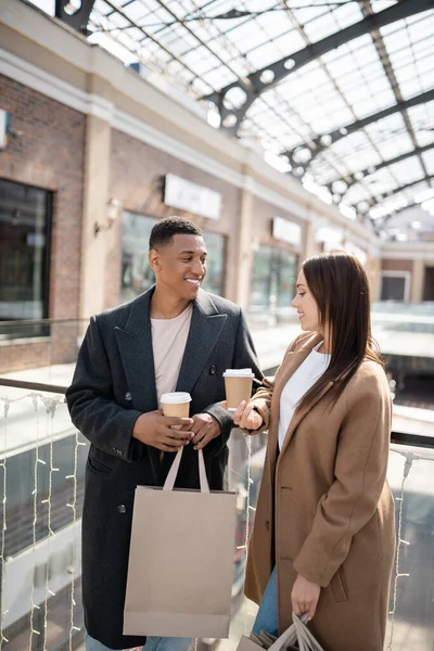 Усміхнена багатоетнічна пара з сумками і кавою, щоб піти розмовляти біля розмитого торгового центру — стокове фото