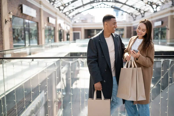 Mujer feliz mirando el teléfono inteligente cerca de novio afroamericano con bolsas de compras - foto de stock