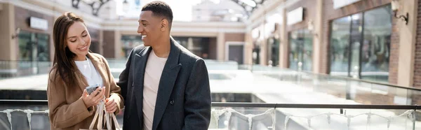 Hombre afroamericano de moda sonriendo cerca de la mujer con el teléfono móvil y bolsas de compras, pancarta - foto de stock