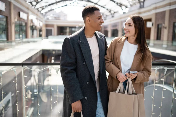 Bonita mujer con teléfono móvil y bolsas de compras sonriendo al hombre afroamericano de moda cerca de centro comercial borrosa - foto de stock