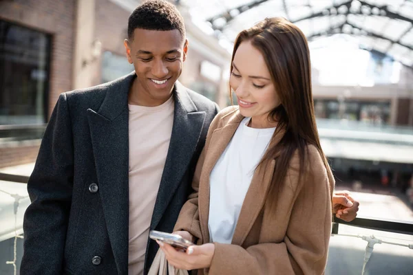 Brunette woman looking at mobile phone near smiling african american boyfriend outdoors — Stock Photo