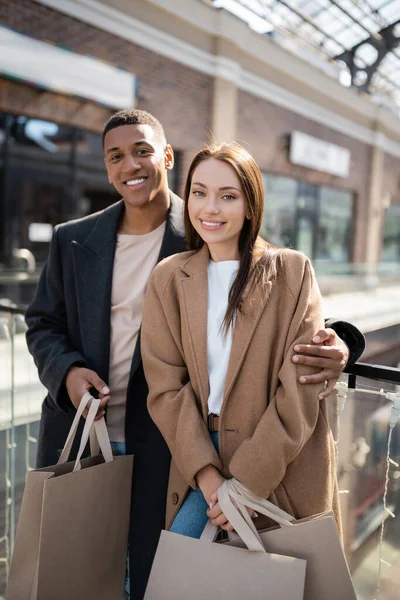 Pleased interracial couple in stylish coats holding shopping bags and looking at camera near mall — Stock Photo