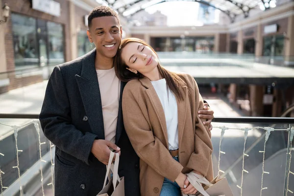 Mujer feliz con los ojos cerrados apoyándose en el novio afroamericano sonriente con bolsas de compras cerca del centro comercial borroso - foto de stock