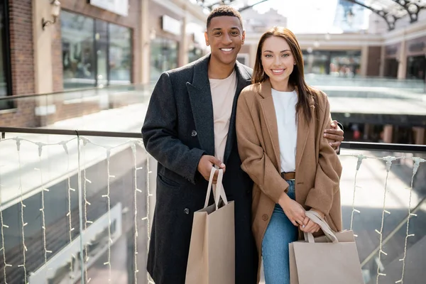 Alegre casal multiétnico em casacos da moda segurando sacos de compras e olhando para a câmera perto de shopping borrado — Fotografia de Stock