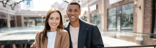 Young brunette woman and african american man in stylish coats smiling at camera on street, banner — Stock Photo